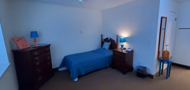 A small bedroom with a single bed covered in a blue bedspread, a wooden headboard, and a pillow. To the left of the bed is a wooden dresser with an orange lamp and a framed picture on top. To the right of the bed is a wooden nightstand with a blue lamp and a telephone. There is a small blue table with a decorative item and a wall-mounted mirror near the door. The room has beige carpet and white walls.