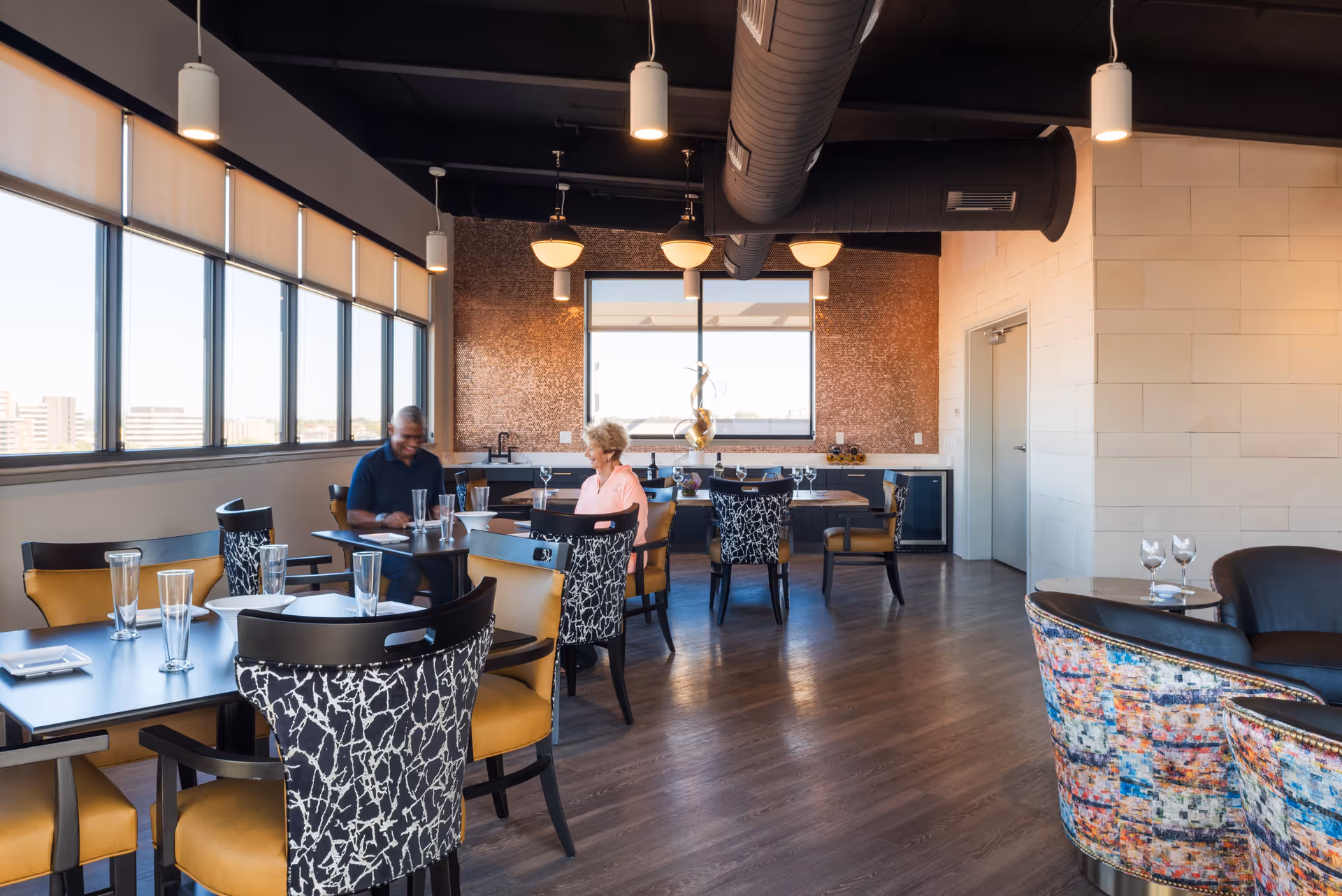 A modern dining room with large windows letting in natural light. Several tables with black tops and chairs with patterned backs and mustard yellow seats are arranged throughout the room. Two people are seated at one of the tables, engaged in conversation. The room features a decorative wall with a window and a sculptural piece, pendant lighting, and a dark exposed ceiling with ductwork.