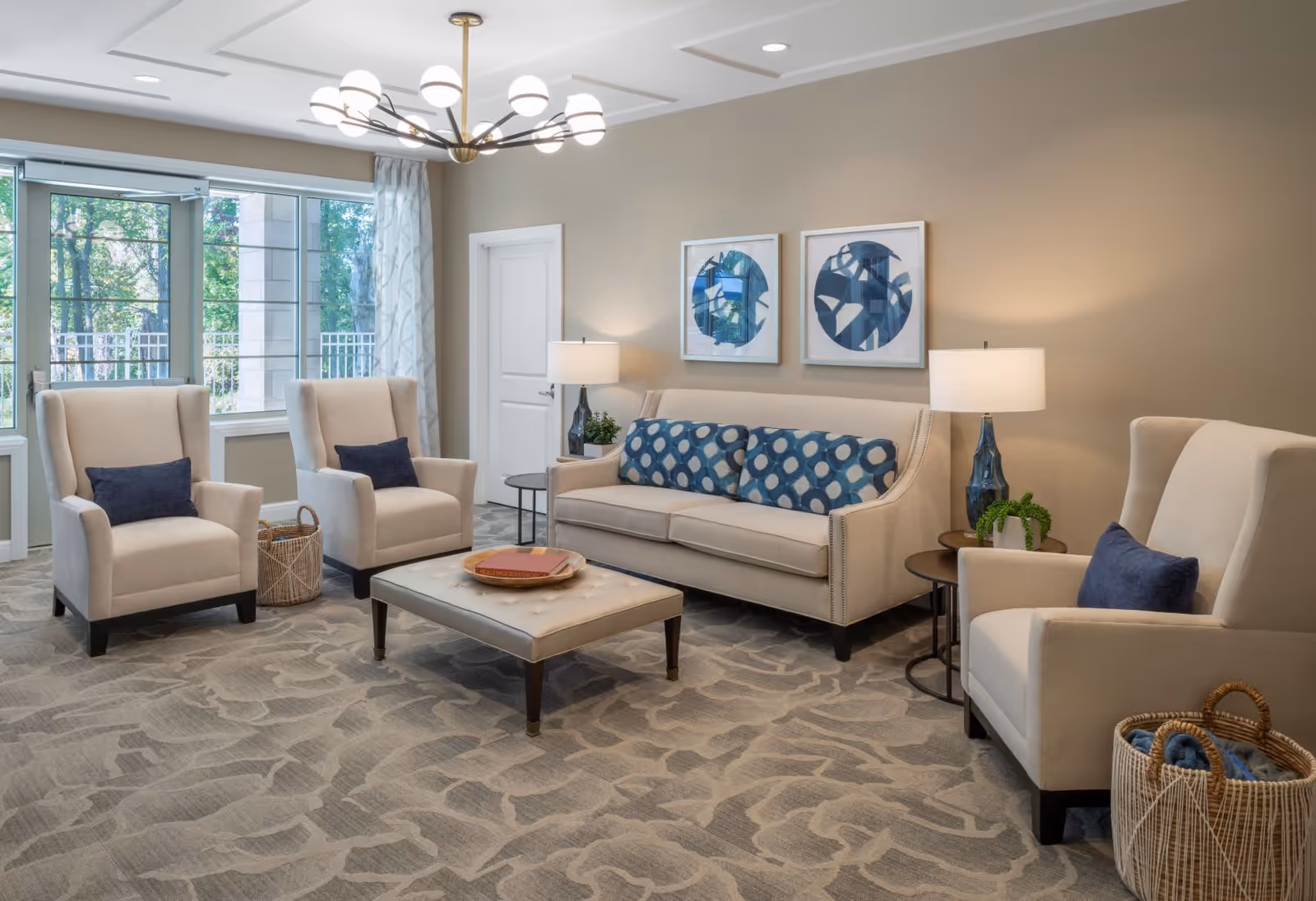 A bright and modern living room with beige walls and carpet featuring a subtle pattern. The room has a beige sofa with three blue patterned cushions, two beige armchairs with dark blue pillows, and a large beige ottoman in the center. Two side tables with lamps and a small plant are placed beside the sofa. Large windows with patterned curtains allow natural light to fill the room, and two abstract blue and white artworks hang on the wall above the sofa. A woven basket with blankets is placed near one of the armchairs.
