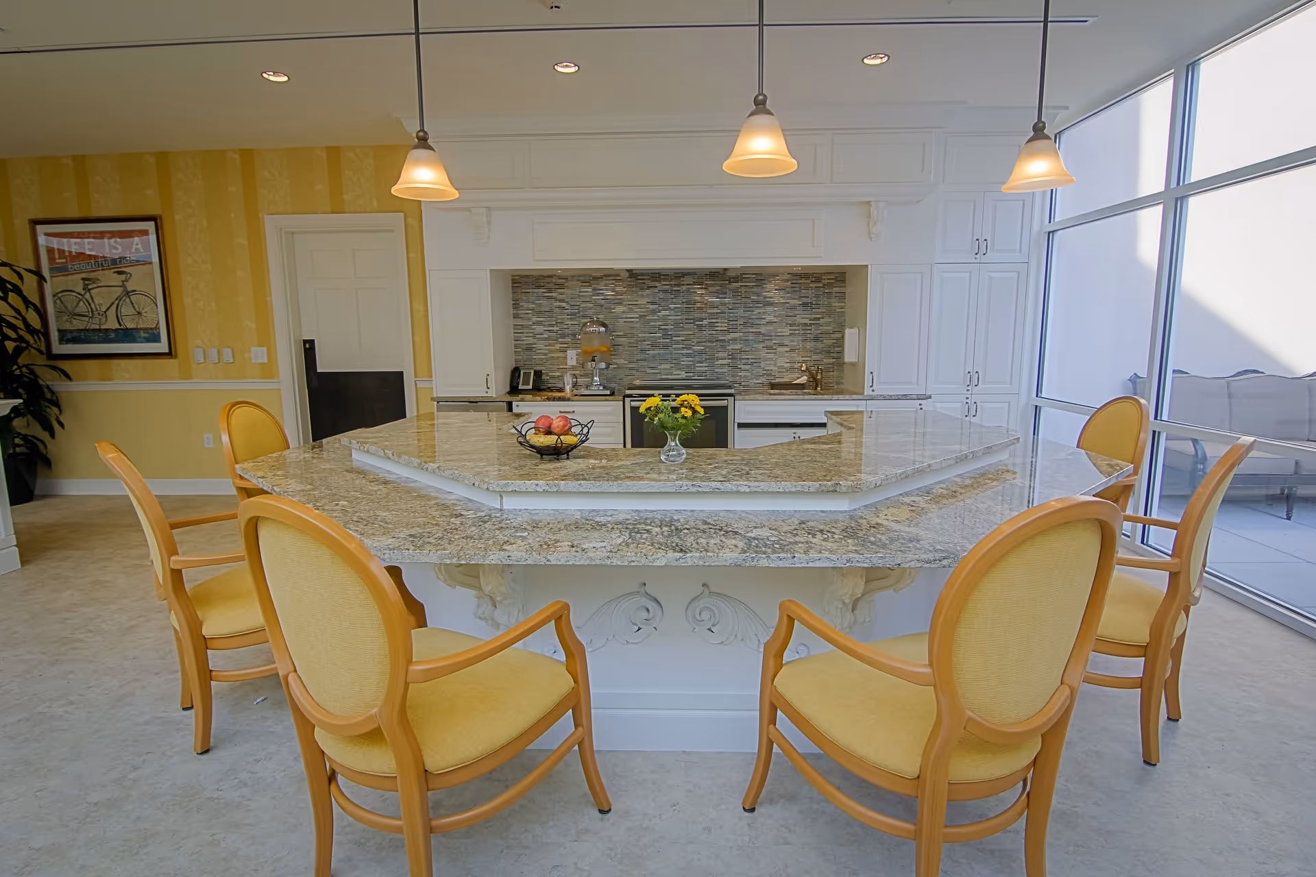 A bright kitchen area with a large granite countertop island surrounded by six wooden chairs with yellow cushions. The kitchen features white cabinetry, a mosaic tile backsplash, and three pendant lights hanging from the ceiling. A glass vase with yellow flowers and a fruit basket are placed on the island. Large windows on the right side let in natural light, and a framed picture with a bicycle and the text 'Life is a beautiful ride' hangs on the yellow wall to the left.