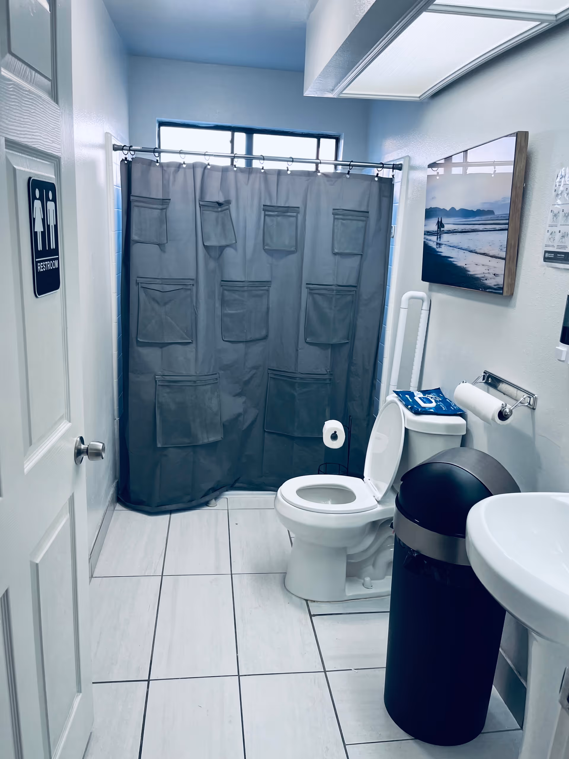A clean bathroom with a white toilet, a black trash can, a white sink, and a shower area with a gray curtain that has multiple pockets. There is a window above the shower and a framed picture of a beach scene on the wall. A restroom sign is visible on the door.