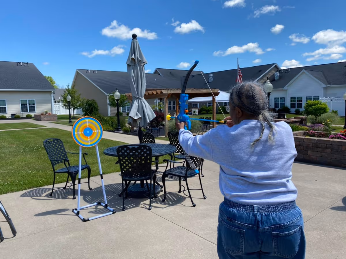 An elderly person with braided hair is outdoors aiming a toy bow and arrow at a colorful target set up on a stand. The scene is in a courtyard with patio furniture, umbrellas, and residential buildings in the background under a blue sky with some clouds.