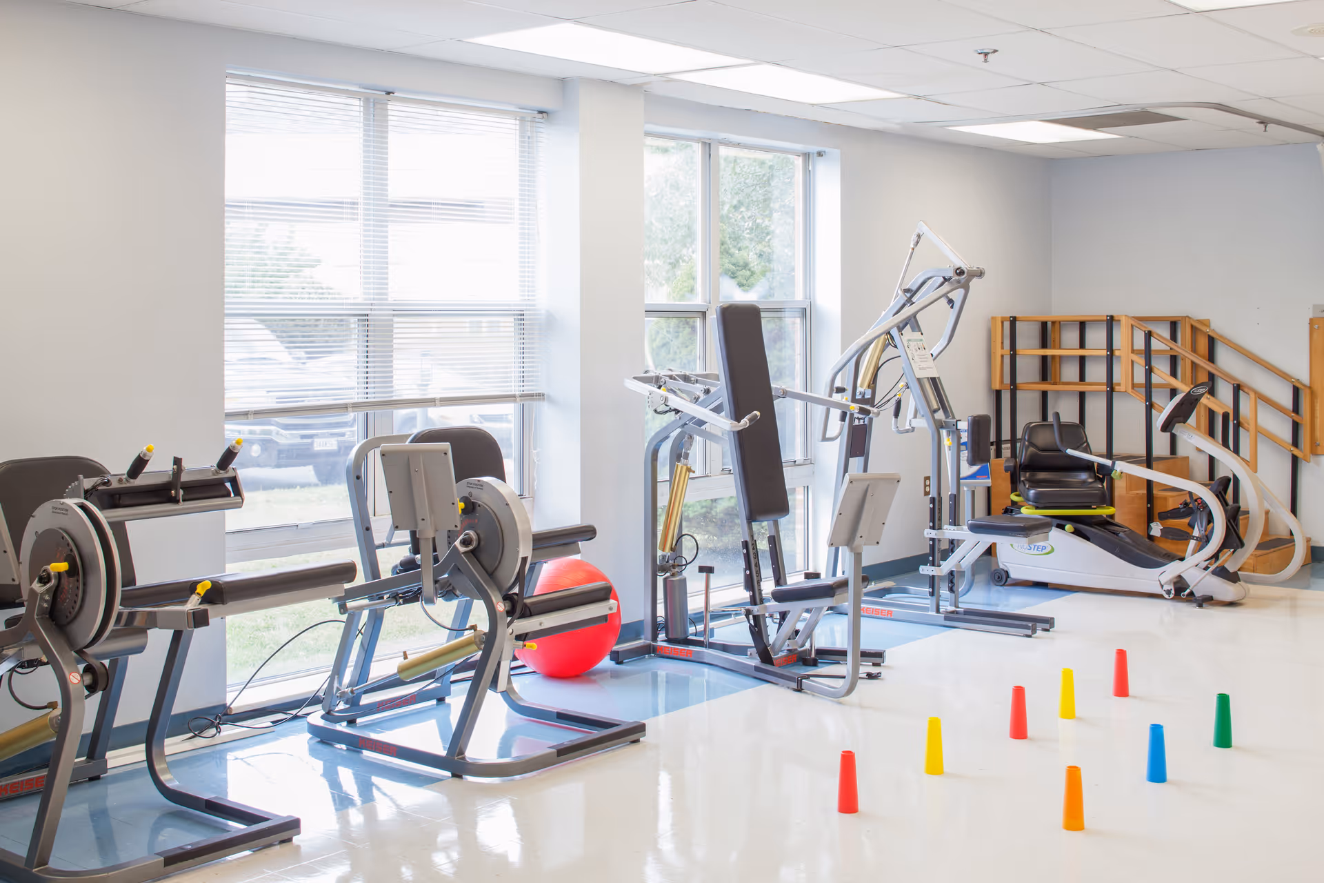 Rehabilitation exercise room with physical therapy equipment, a red exercise ball, and colored cones by large windows.
