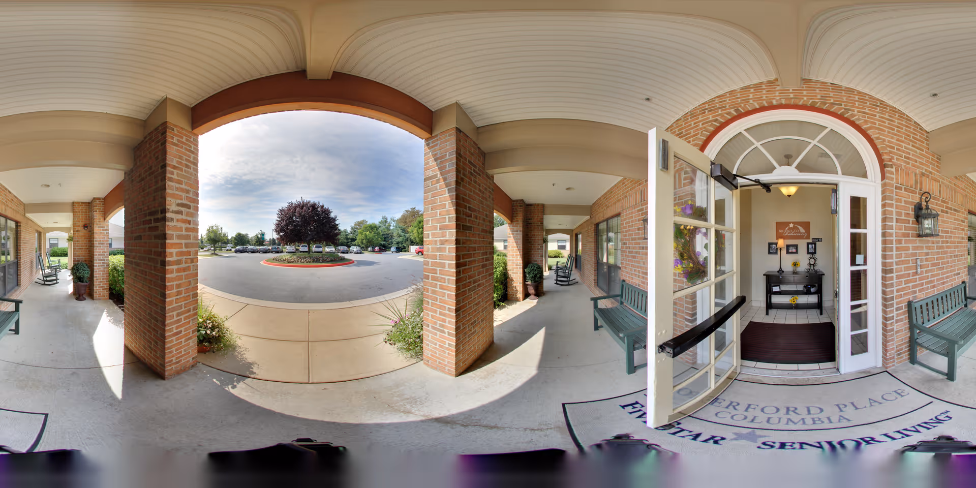 Covered brick entrance with benches and an open glass door leading into a senior living facility and a circular driveway with a landscaped island.