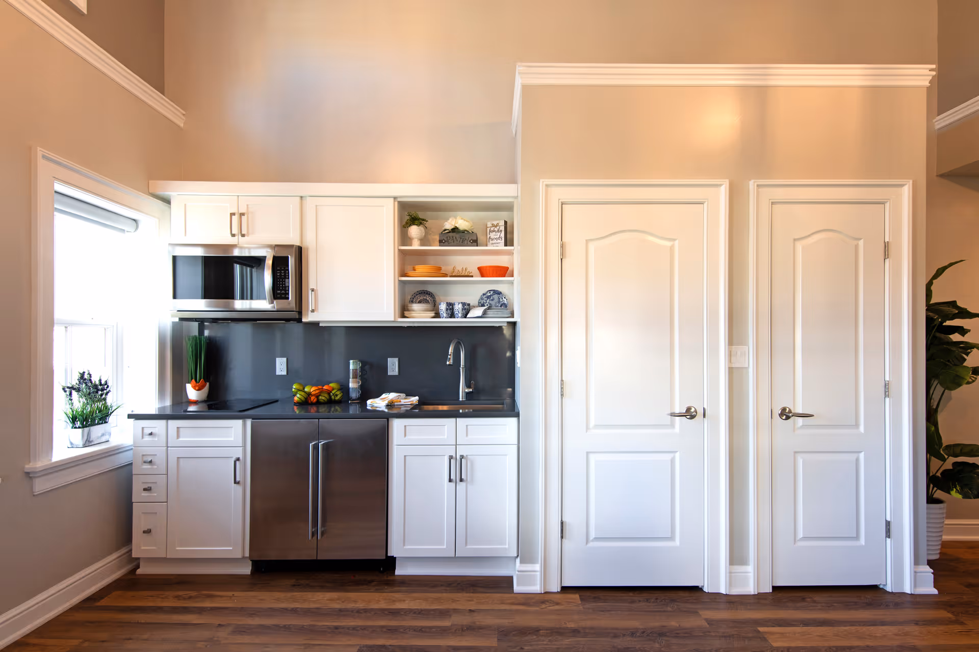 A modern kitchen area with white cabinets, a black countertop, and stainless steel appliances including a microwave and a dishwasher. There is a window with a potted plant on the left side, open shelves with dishes and decorative items above the sink, and two white doors on the right side. The floor is wooden and the walls are painted beige.