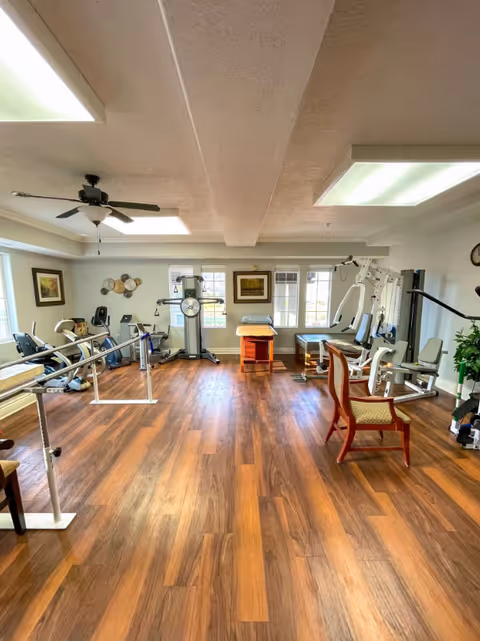 A spacious senior living fitness room with wooden flooring, various exercise equipment including a stationary bike, weight machines, parallel bars, and a ceiling fan. The room is well-lit with large windows and fluorescent ceiling lights, decorated with framed pictures and a potted plant.