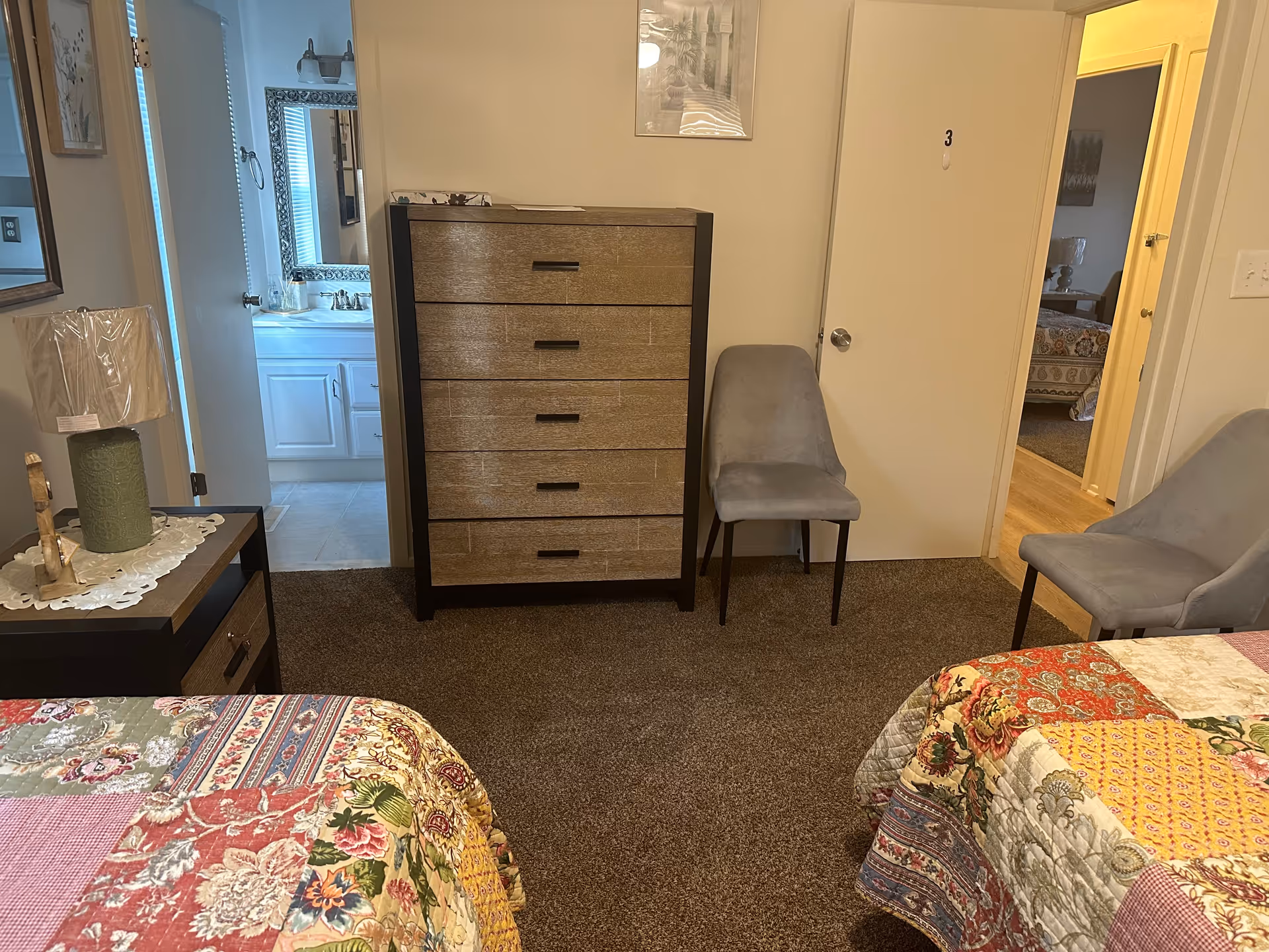 Interior view of a bedroom in an assisted living facility showing two beds with colorful patchwork quilts, a wooden dresser with five drawers, two gray upholstered chairs, a nightstand with a green lamp, and a doorway leading to a bathroom with a sink and mirror.