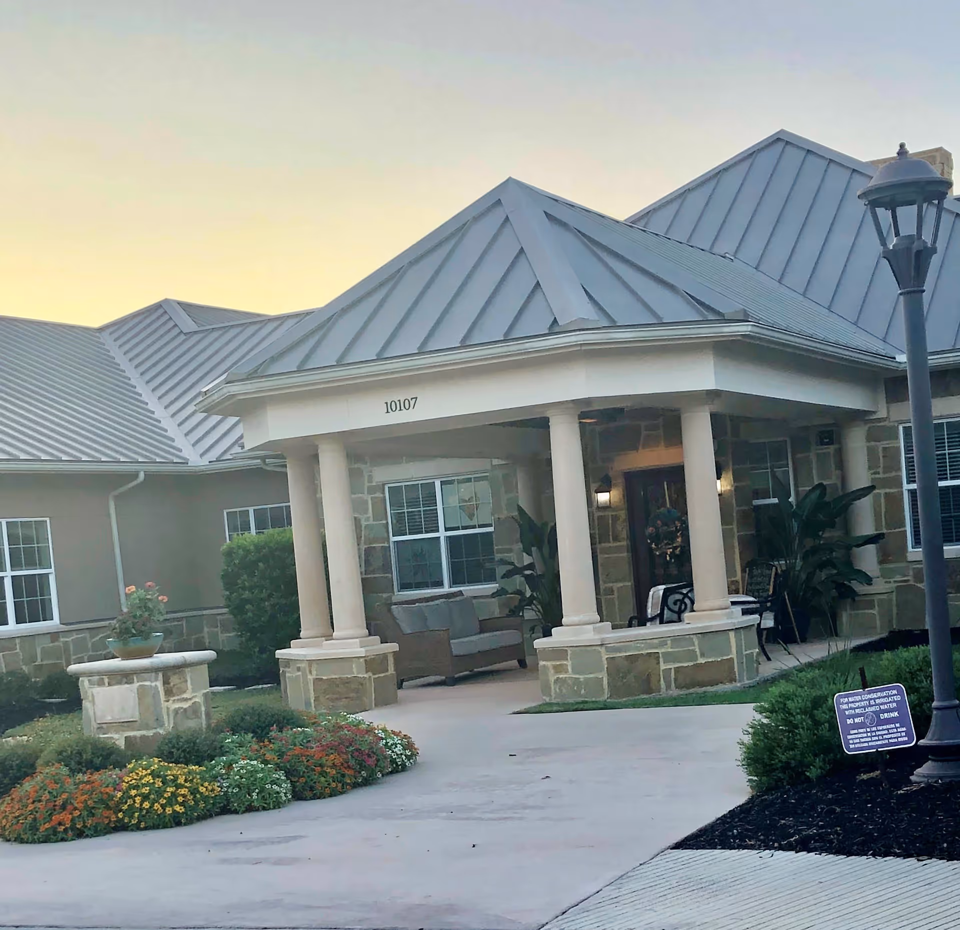 Exterior view of a senior living facility entrance with a covered porch supported by columns, stone and beige walls, metal roof, outdoor seating, landscaped flower beds, and a street lamp near the walkway.