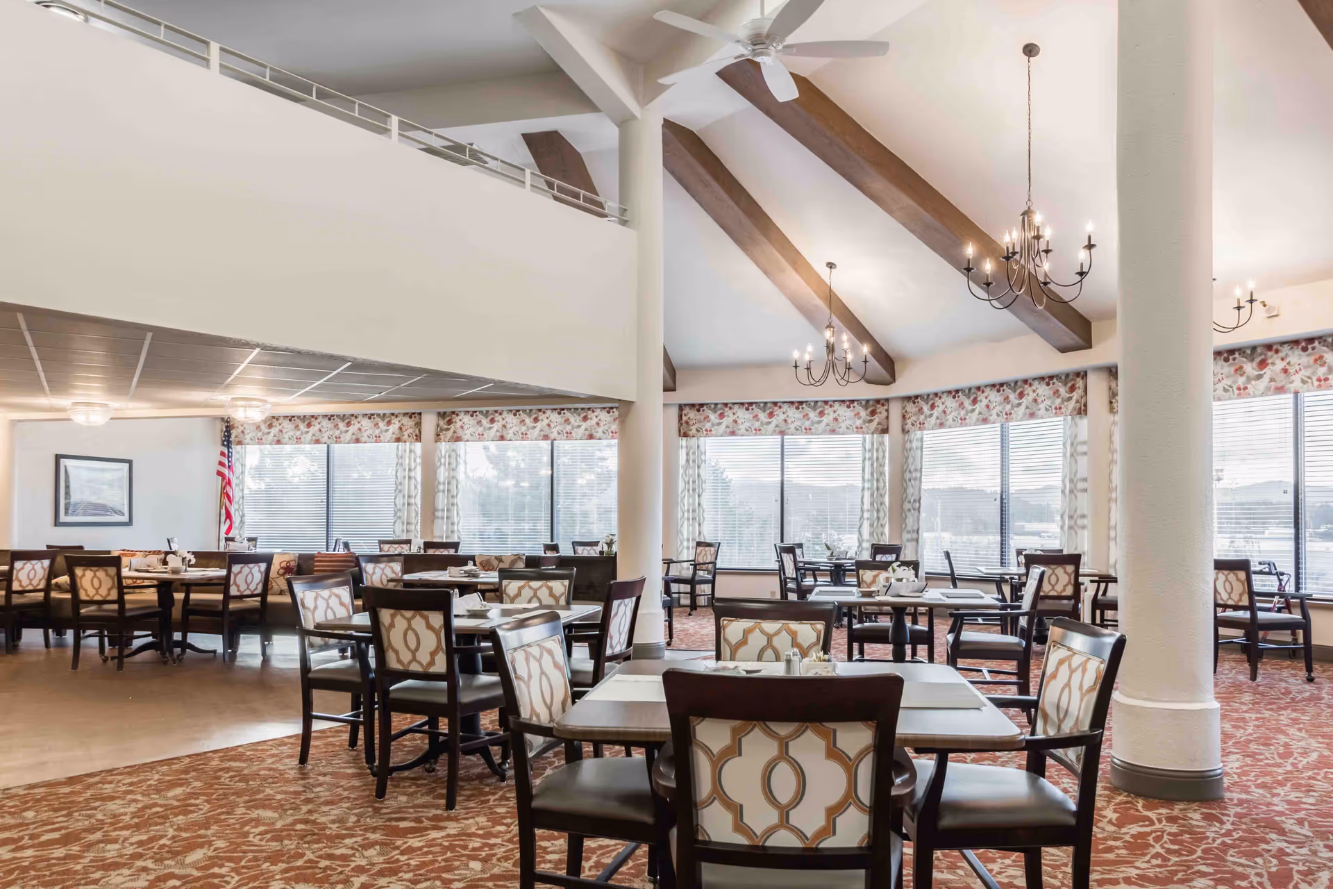 A spacious dining room with multiple tables and chairs arranged neatly. The room features large windows with floral valances and blinds, allowing natural light to fill the space. The ceiling has exposed wooden beams and several chandeliers providing warm lighting. The floor is carpeted with a patterned design, and there are white columns supporting the ceiling.