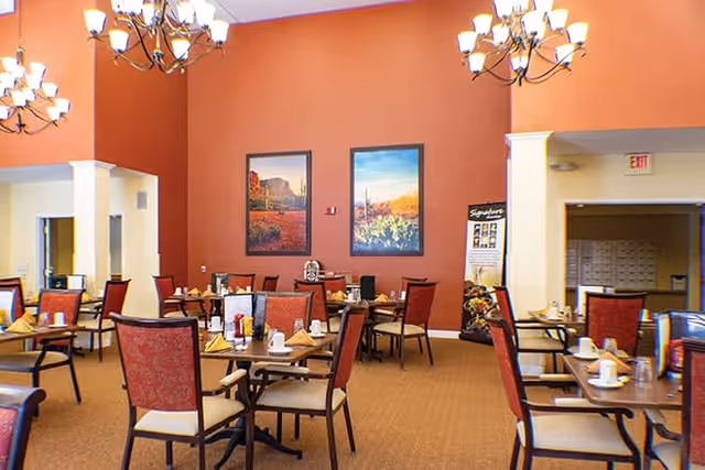 Dining room with multiple tables and chairs arranged neatly. Each table is set with cups, napkins, and condiments. The walls are painted a warm orange color and decorated with two landscape paintings. Elegant chandeliers hang from the ceiling providing soft lighting.