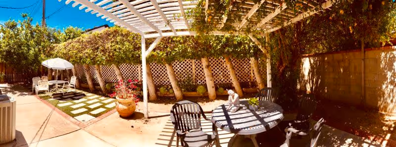 A sunny outdoor patio area with a white pergola casting shadows over a round table and several chairs. There is a small statue on the table and a large potted plant nearby. In the background, there is a lattice fence with trees and bushes providing greenery. To the left, there is a small seating area with a white umbrella and chairs on a patch of grass with stone pavers.