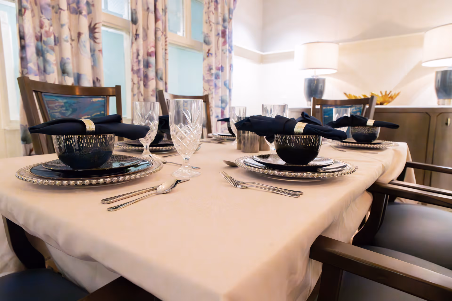 A dining table set for four with black bowls, black napkins with gold napkin rings, clear crystal glasses, and silverware on a beige tablecloth. The background shows floral curtains and two table lamps on a sideboard.