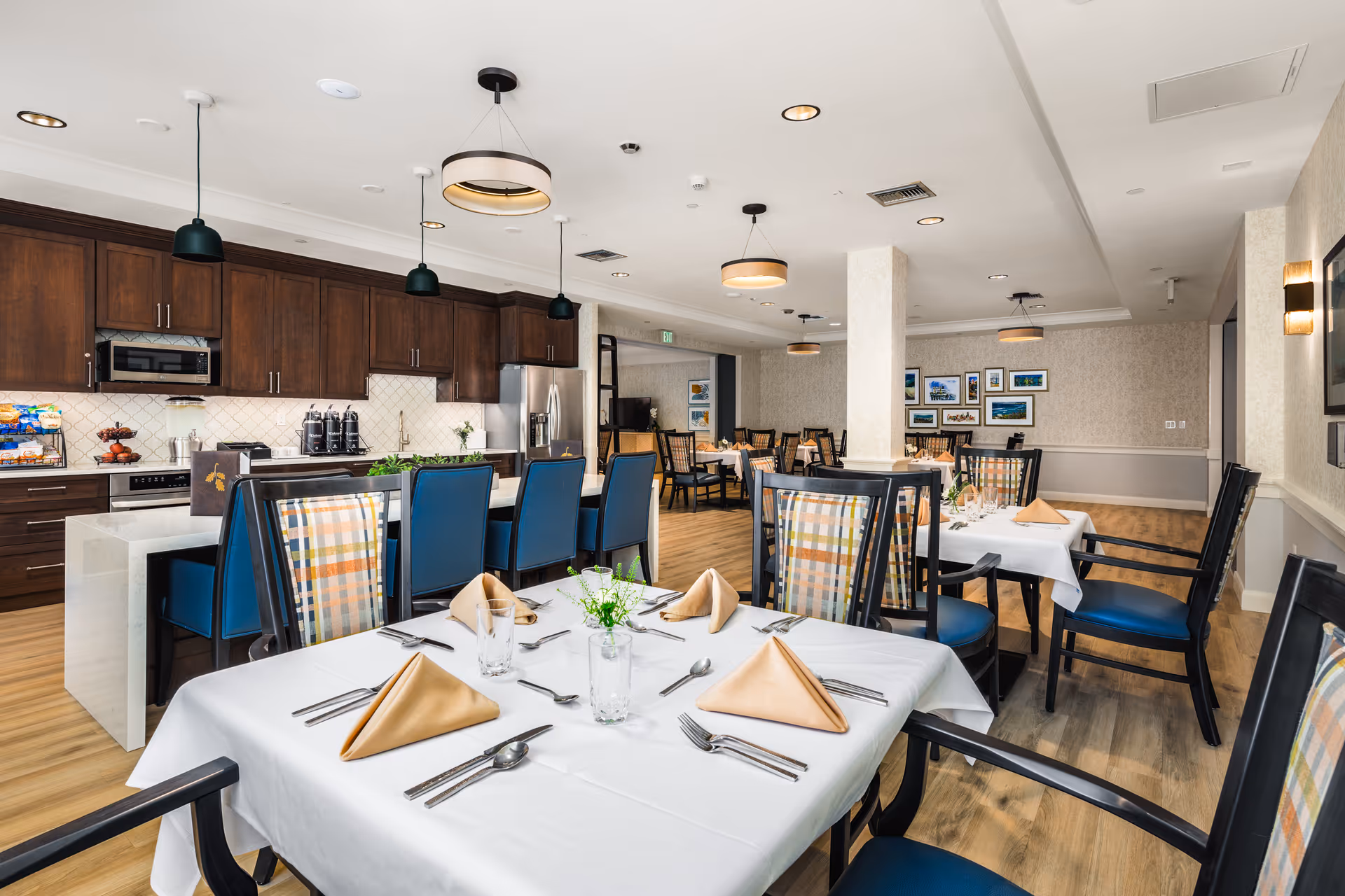 A bright and spacious dining room with several tables covered in white tablecloths, each set with beige folded napkins, silverware, and glasses. The chairs have dark frames with blue seats and plaid backs. In the background, there is a kitchen area with dark wooden cabinets, a stainless steel refrigerator, a microwave, and a countertop with snacks and beverage dispensers. The room has modern pendant and ceiling lights, light wood flooring, and framed artwork on the walls.