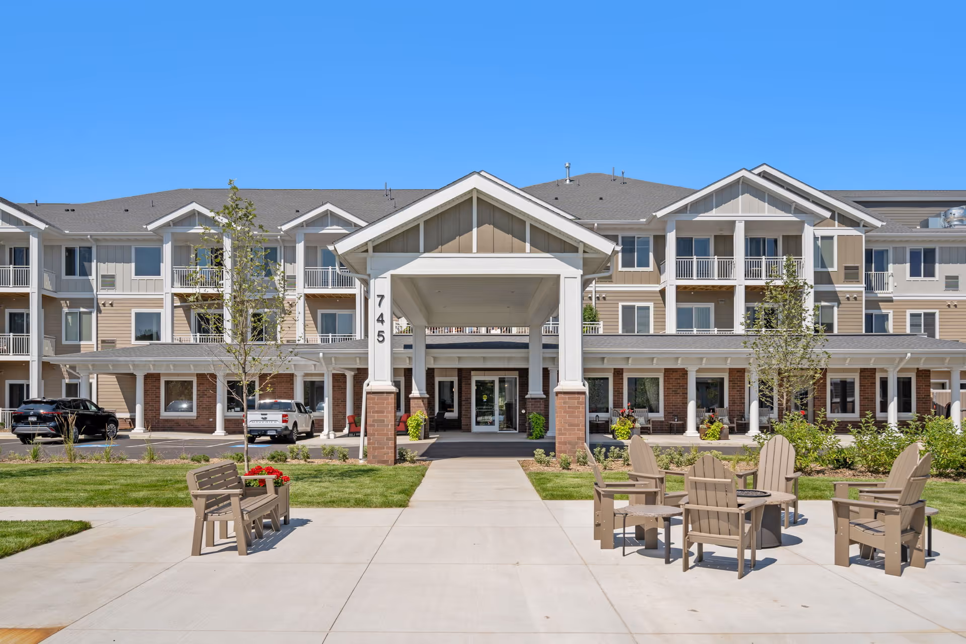 Front exterior view of The Sycamore of River Falls senior living facility with a covered entrance, multiple balconies, parked cars, and outdoor seating area with chairs and a fire pit under a clear blue sky.