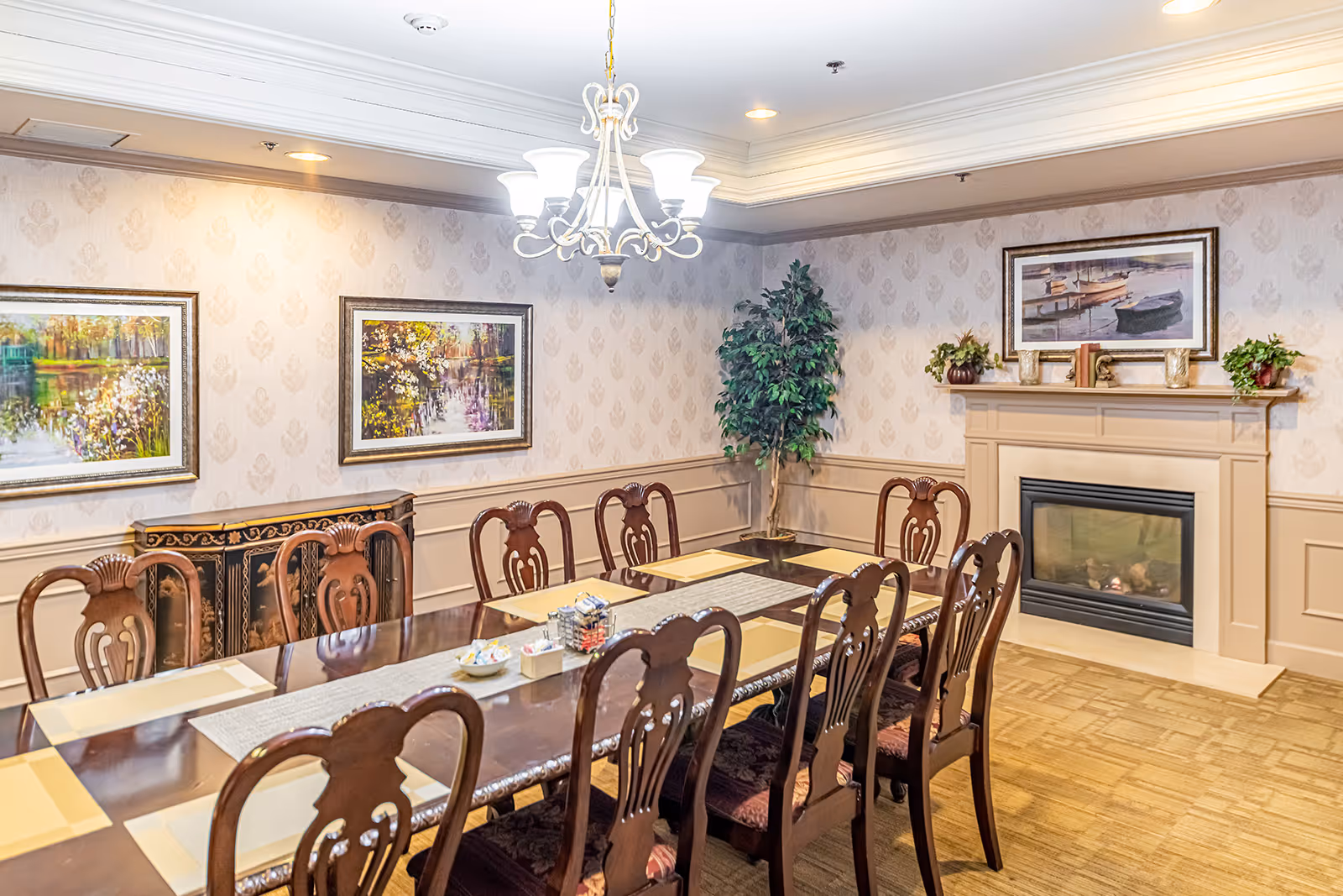 A formal dining room with a long wooden table surrounded by eight ornate wooden chairs. The table is set with placemats and a centerpiece. The room features a beige patterned wallpaper, a chandelier hanging from the ceiling, framed landscape paintings on the walls, a fireplace with decorative items on the mantel, and a potted plant in the corner.