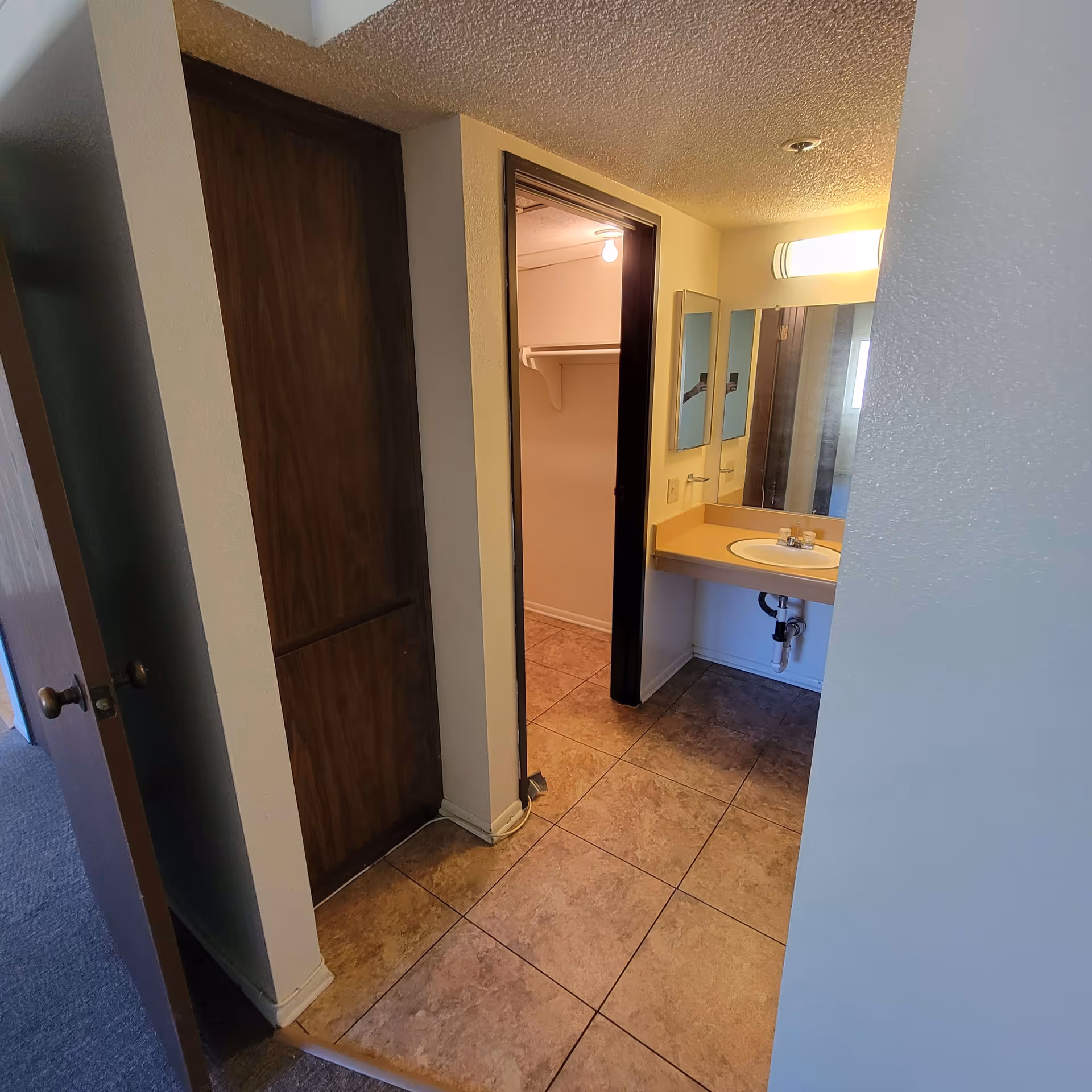 Interior view of a small bathroom area in Briarwood Manor featuring a sink with a mirror and light above it, a tiled floor, a wooden door to the left, and an open doorway leading to a closet with a light bulb and shelf inside.