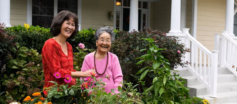 Two elderly women smiling and tending to a garden with colorful flowers in front of a house with white railings and steps.