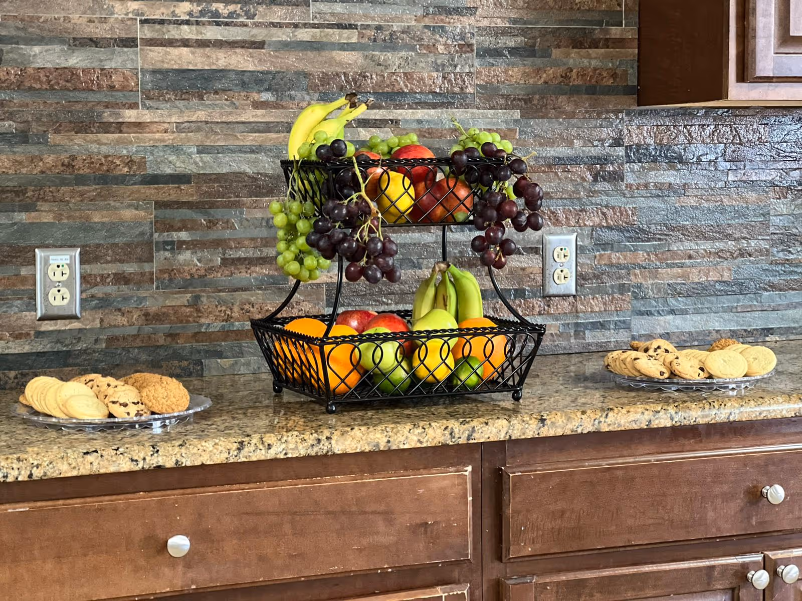 A kitchen countertop with a two-tiered black wire fruit basket filled with bananas, grapes, apples, oranges, and pears. On either side of the fruit basket are plates with assorted cookies. The backsplash is made of multi-colored stone tiles, and there are two electrical outlets visible on the wall. Below the countertop are wooden cabinets with silver knobs.