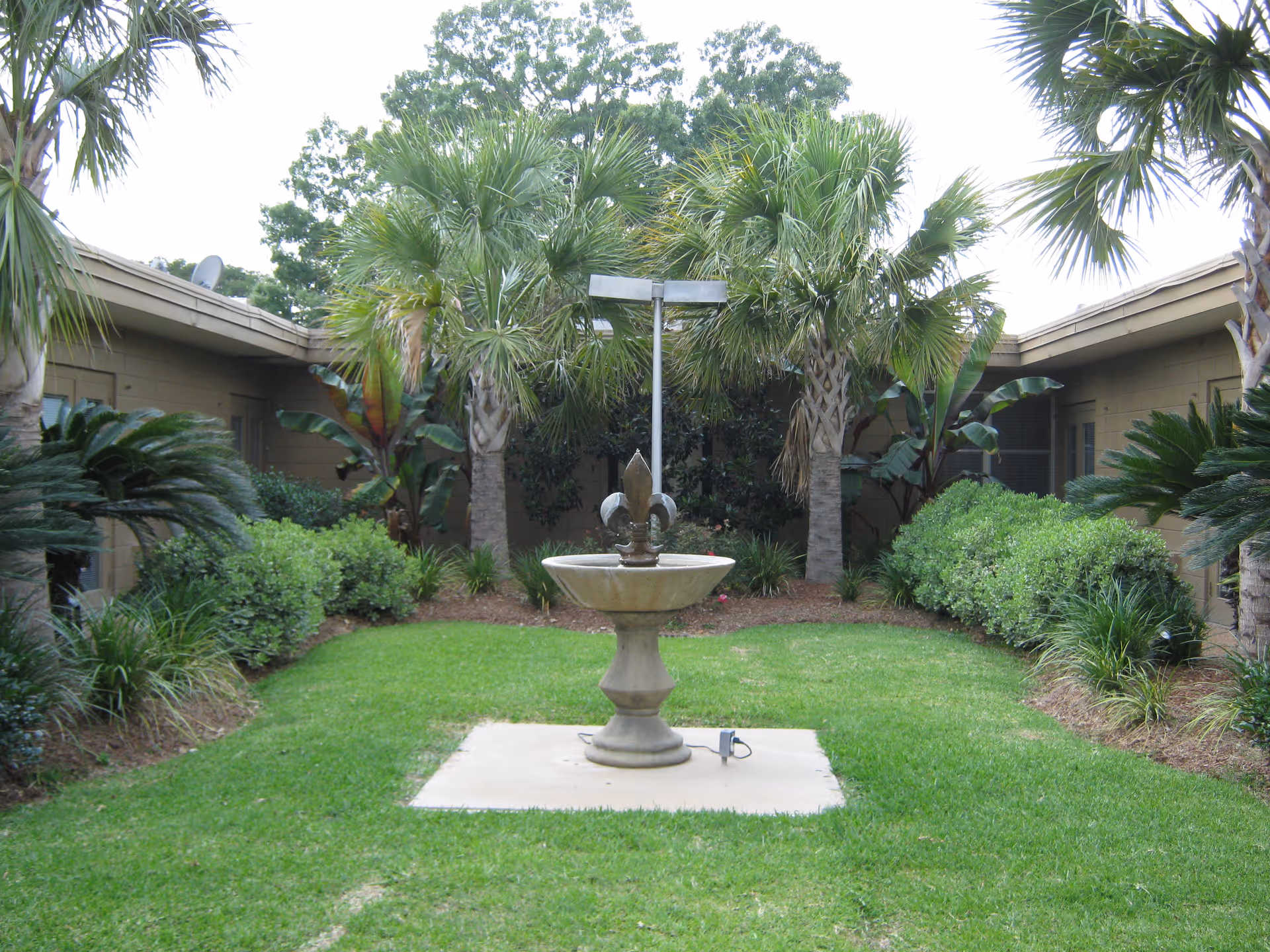 A landscaped courtyard with a central stone fountain surrounded by palm trees and trimmed bushes between single-story building wings.