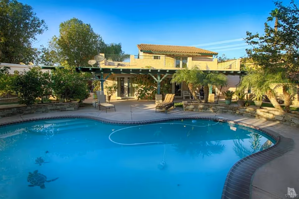 Outdoor swimming pool with a curved shape and turtle designs at the bottom, surrounded by a patio area with lounge chairs and greenery including trees and potted plants. In the background, there is a two-story building with a balcony and a pergola-covered patio area.