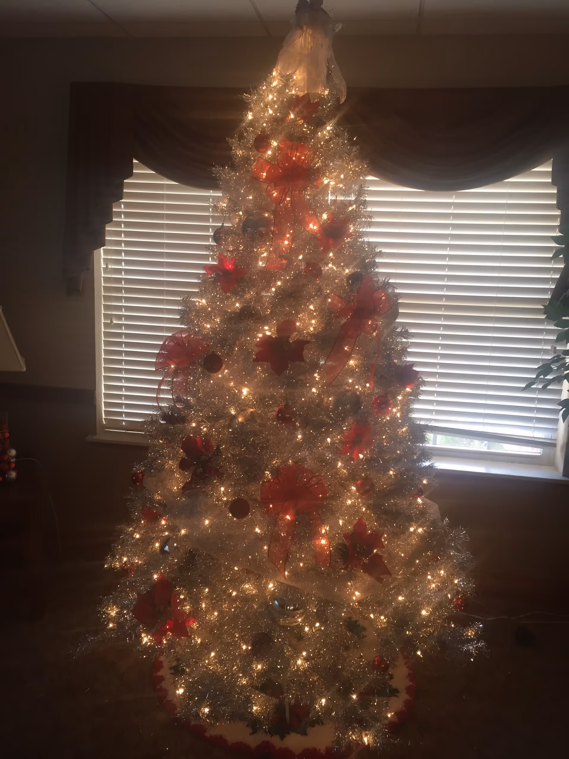 A brightly lit Christmas tree decorated with red bows, ornaments, and white lights stands in front of a window with closed blinds and dark curtains in a cozy room.