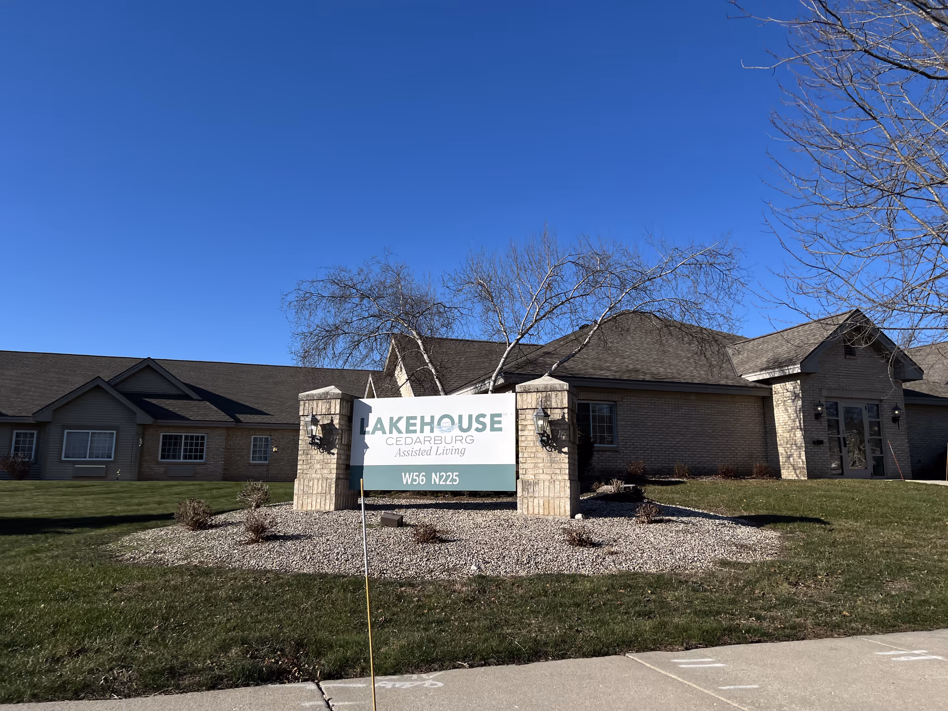 Exterior view of LakeHouse Cedarburg Assisted Living facility with a clear blue sky, a large sign in front displaying the facility name and address, surrounded by grass, small bushes, and a paved sidewalk.