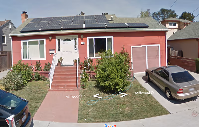 Front exterior view of a single-story house painted in reddish color with a white door and windows. The house has solar panels installed on the roof. There are steps leading up to the front door with railings on both sides. A driveway on the right side has a parked silver car. The front yard has green grass, bushes, and a garden hose lying on the lawn. The street name Millwood Dr is visible on the pavement.