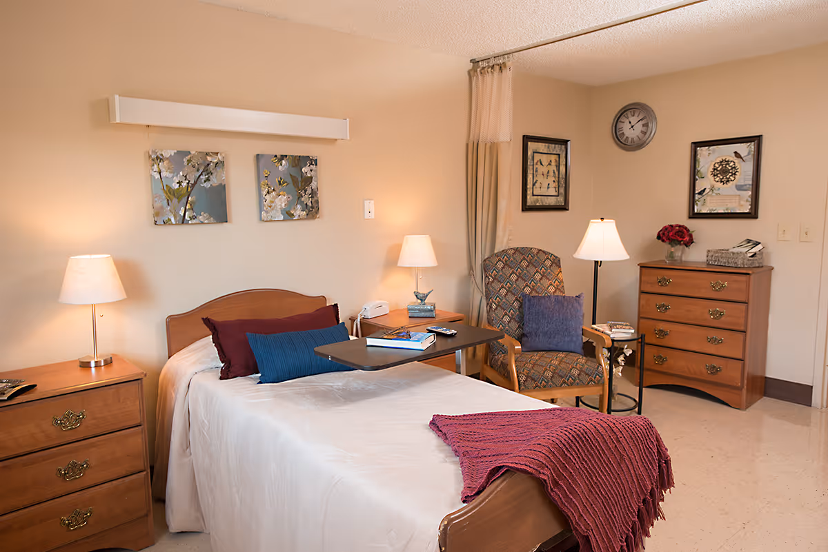 A cozy bedroom in a healthcare facility featuring a single bed with white bedding, a burgundy throw blanket, and decorative pillows. There are two wooden nightstands with lamps on either side of the bed. A patterned armchair with a blue cushion is positioned next to a wooden dresser with a vase of red flowers and framed artwork on the walls. A clock is mounted on the wall above the dresser.