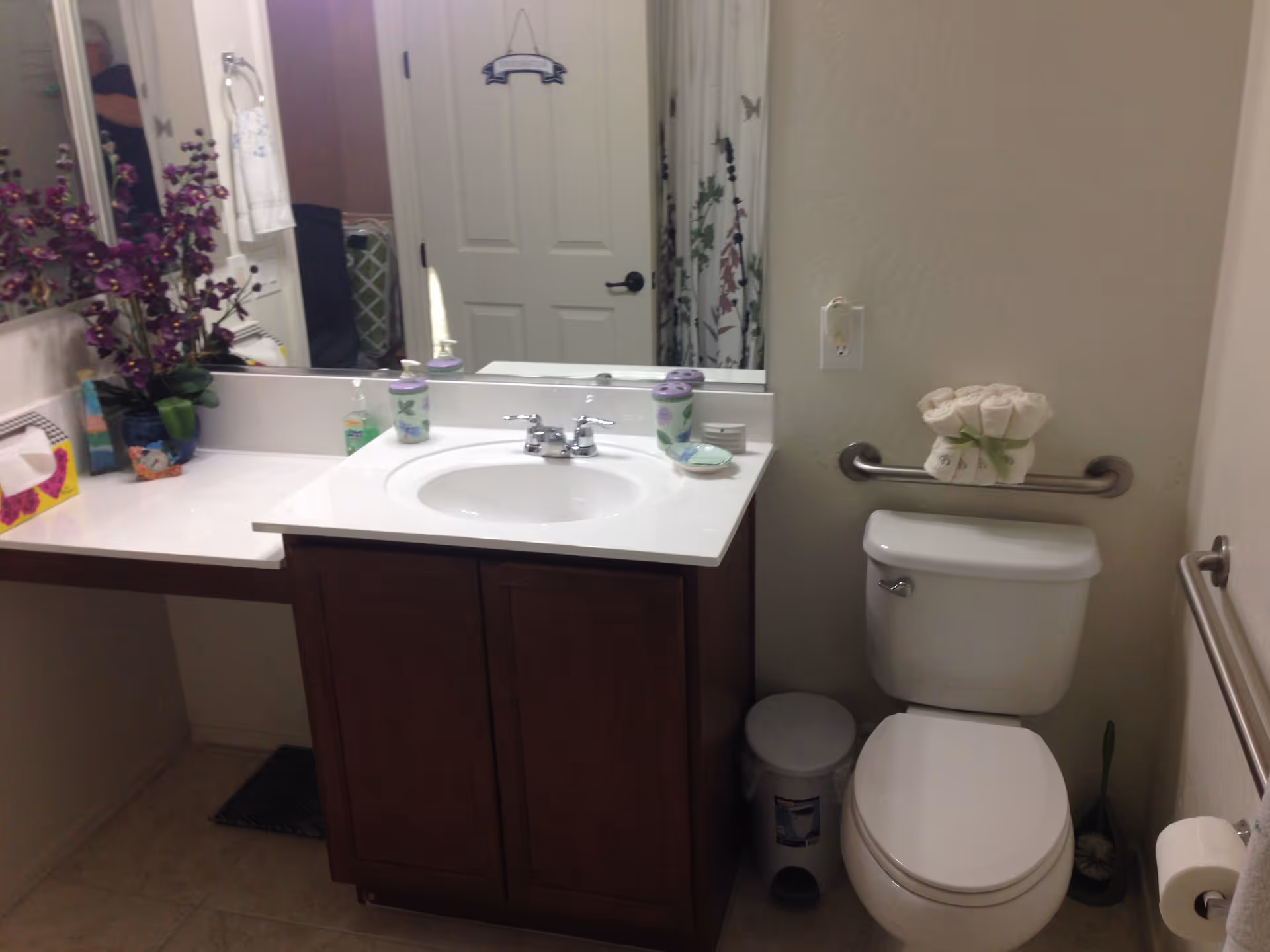 A bathroom with a white sink and countertop, a wooden cabinet below, a toilet with a grab bar behind it, and a trash can next to the toilet. There are decorative items including a purple flower arrangement, soap dispenser, and a small dish on the counter. A mirror is above the sink reflecting a door and a shower curtain with a floral pattern.