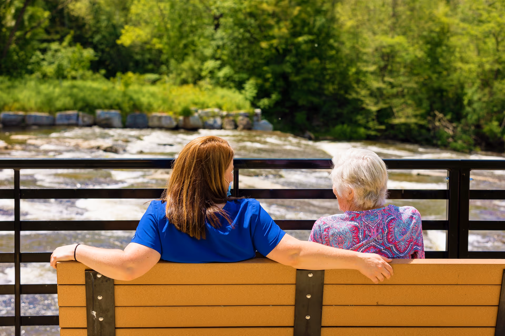 A younger woman in a blue shirt with a face mask pulled down sits on a bench with her arm around an elderly woman wearing a colorful patterned shirt. They are outdoors, facing a flowing river with green trees in the background.