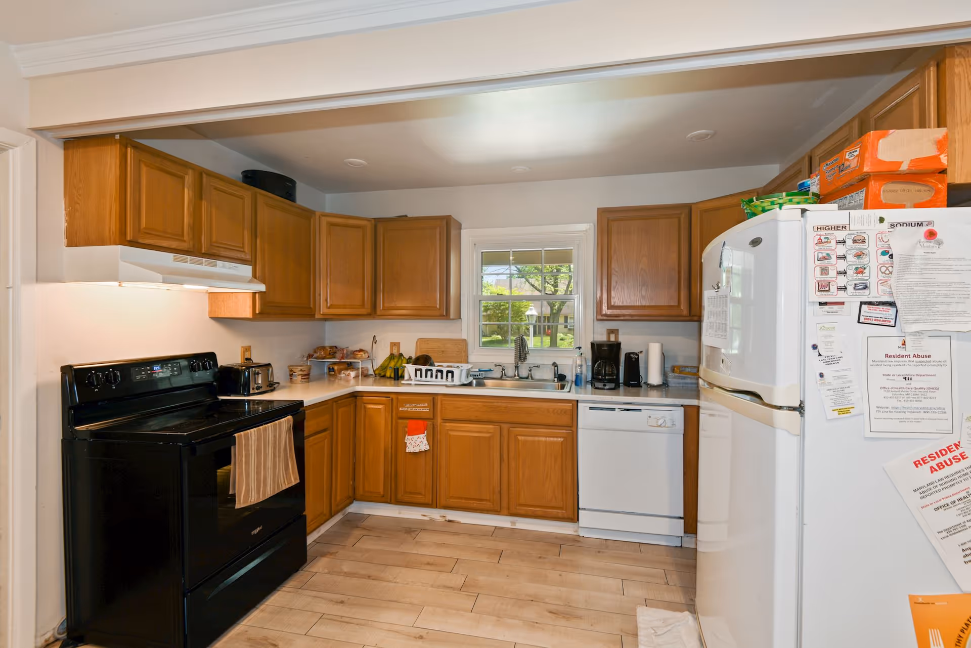 A kitchen with wooden cabinets, a black stove with a towel hanging on the handle, a white dishwasher, and a white refrigerator covered with various papers and notices. There is a window above the sink showing a green outdoor area. The countertop has a toaster, bananas, a coffee maker, and other kitchen items.