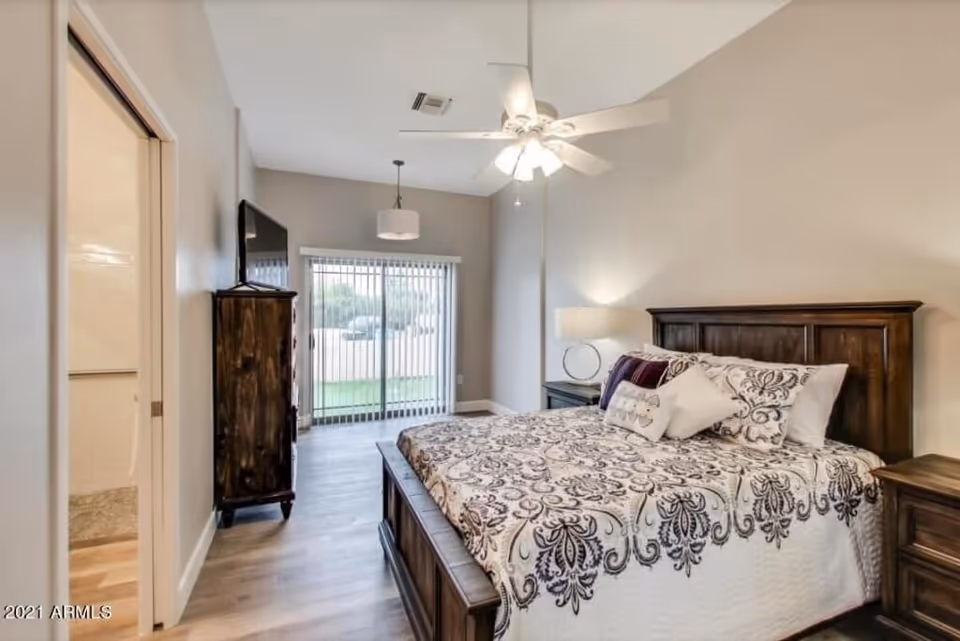 A bedroom with a large wooden bed featuring a patterned bedspread and multiple pillows. There is a wooden nightstand with a lamp on the right side of the bed. A wooden dresser with a flat-screen TV on top is positioned against the left wall. Sliding glass doors with vertical blinds lead to an outdoor area. The room has light-colored walls, a ceiling fan with lights, and wood flooring. A doorway on the left side leads to a bathroom.