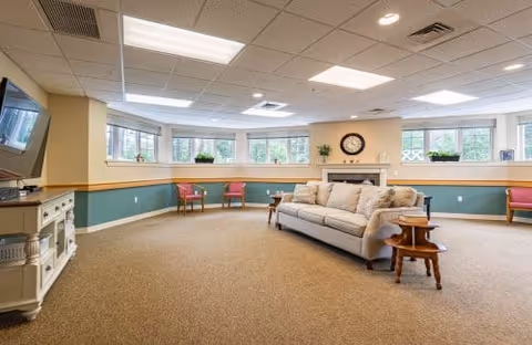 Bright communal living room with a sofa, side tables, fireplace and TV beneath a row of bay windows.
