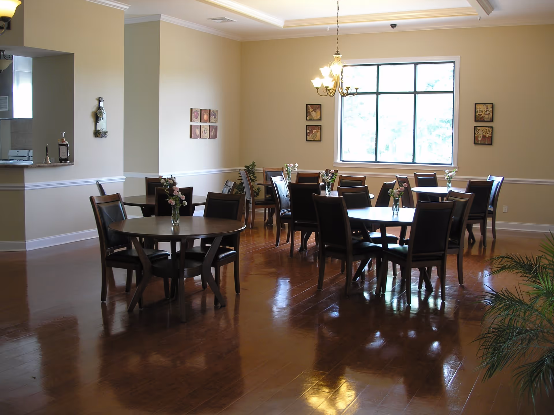 Bright dining room with several round tables and chairs, small floral centerpieces, a chandelier, and a large window.