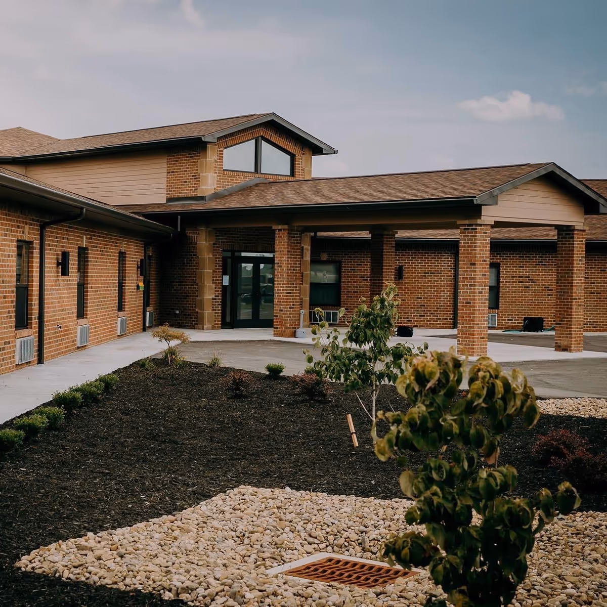 Covered entrance and portico of a brick assisted-living building with landscaped beds and a stone drainage area in front.