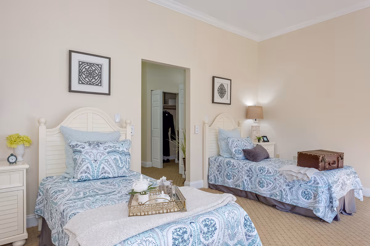 A bedroom with two twin beds featuring matching blue and white patterned bedding. Each bed has multiple pillows and a throw blanket. There is a nightstand with a lamp and a small clock on the left side of the left bed, and a lamp and a small plant on the nightstand next to the right bed. A brown suitcase is placed on the right bed. The walls are light beige with two framed decorative artworks above each bed. An open doorway leads to a closet with white double doors and some hanging clothes inside.