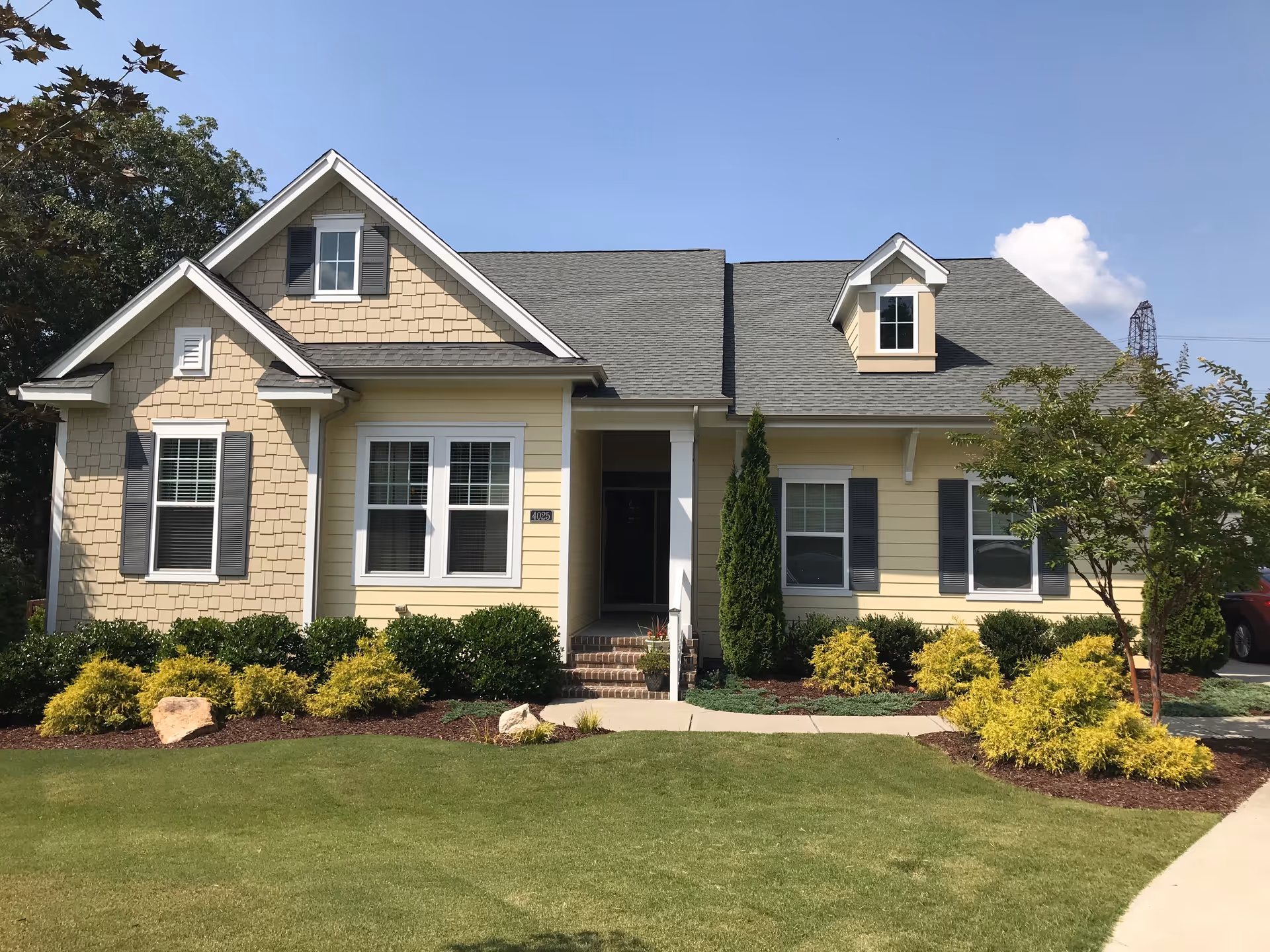 Front exterior view of a single-story house with beige siding, gray roof shingles, white window frames, and dark shutters. The house has a small covered porch with steps leading up to the front door. There are well-maintained bushes, shrubs, and a tree in the landscaped front yard with a green lawn and a clear blue sky in the background.