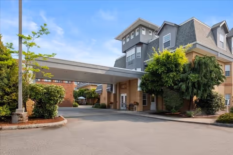 Exterior view of a multi-story senior living facility building with a covered driveway entrance, surrounded by trees and shrubs under a clear blue sky.