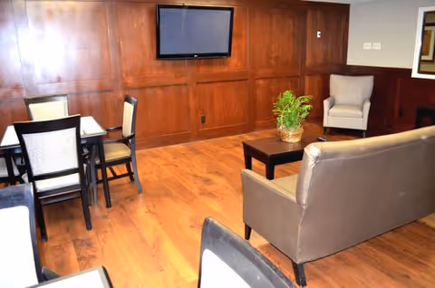 A cozy interior room with wooden flooring and wood-paneled walls featuring a mounted flat-screen TV. The room contains a brown leather sofa, a small coffee table with a potted plant, a beige armchair, and a dining table with four chairs.