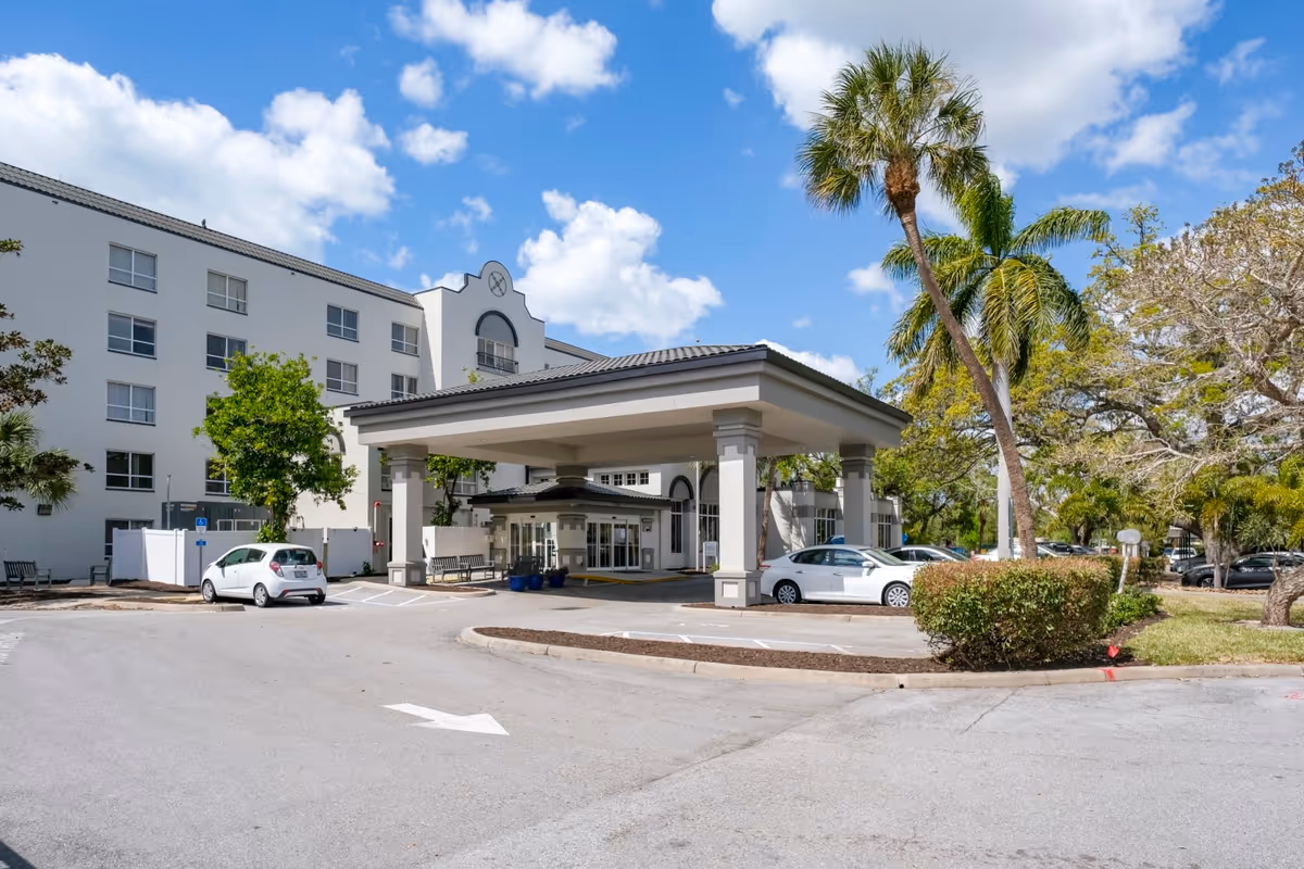 Exterior view of the Grand Villa of Sarasota building entrance with a covered drop-off area, several parked cars, palm trees, and a partly cloudy blue sky.