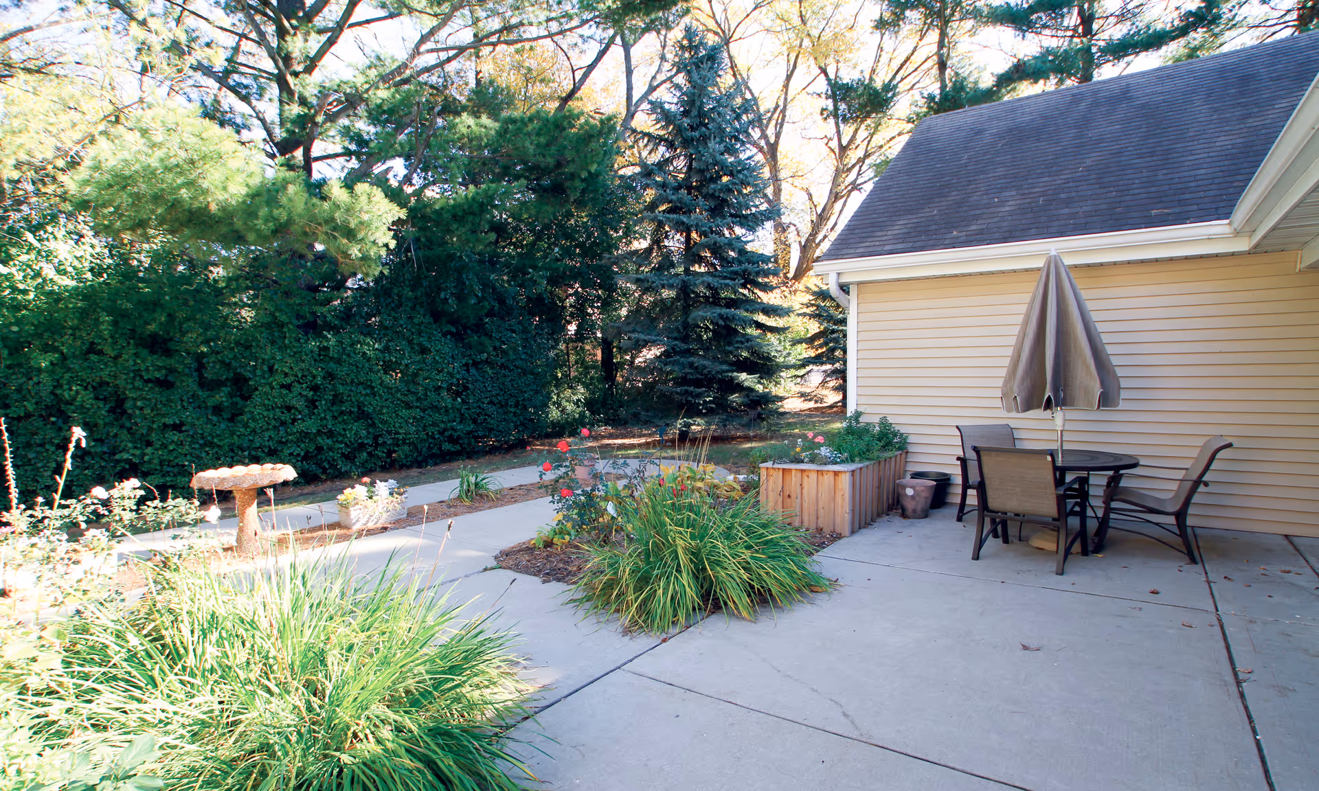 Sunlit outdoor patio with a table, chairs and umbrella next to a building, surrounded by planters, walkways and trees.