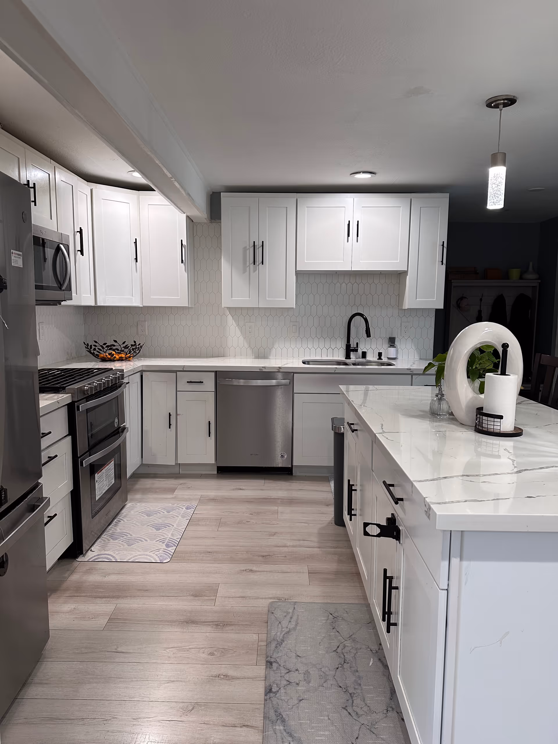 Modern kitchen with white cabinets, stainless steel appliances including a refrigerator, oven, microwave, and dishwasher. The kitchen features a white marble countertop island with a decorative white circular sculpture, a small plant, and a paper towel holder. The backsplash has a white hexagonal tile design, and the floor is light wood. There are two rugs on the floor, one near the oven and one near the island. A pendant light hangs from the ceiling.