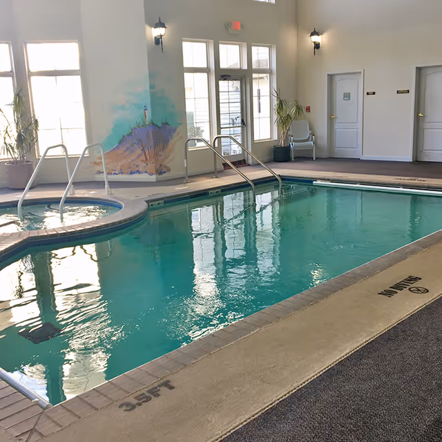 Indoor swimming pool area with clear water, a hot tub adjacent to the pool, metal handrails for entry, large windows letting in natural light, a mural of a lighthouse on the wall, potted plants, and a chair near two closed doors.