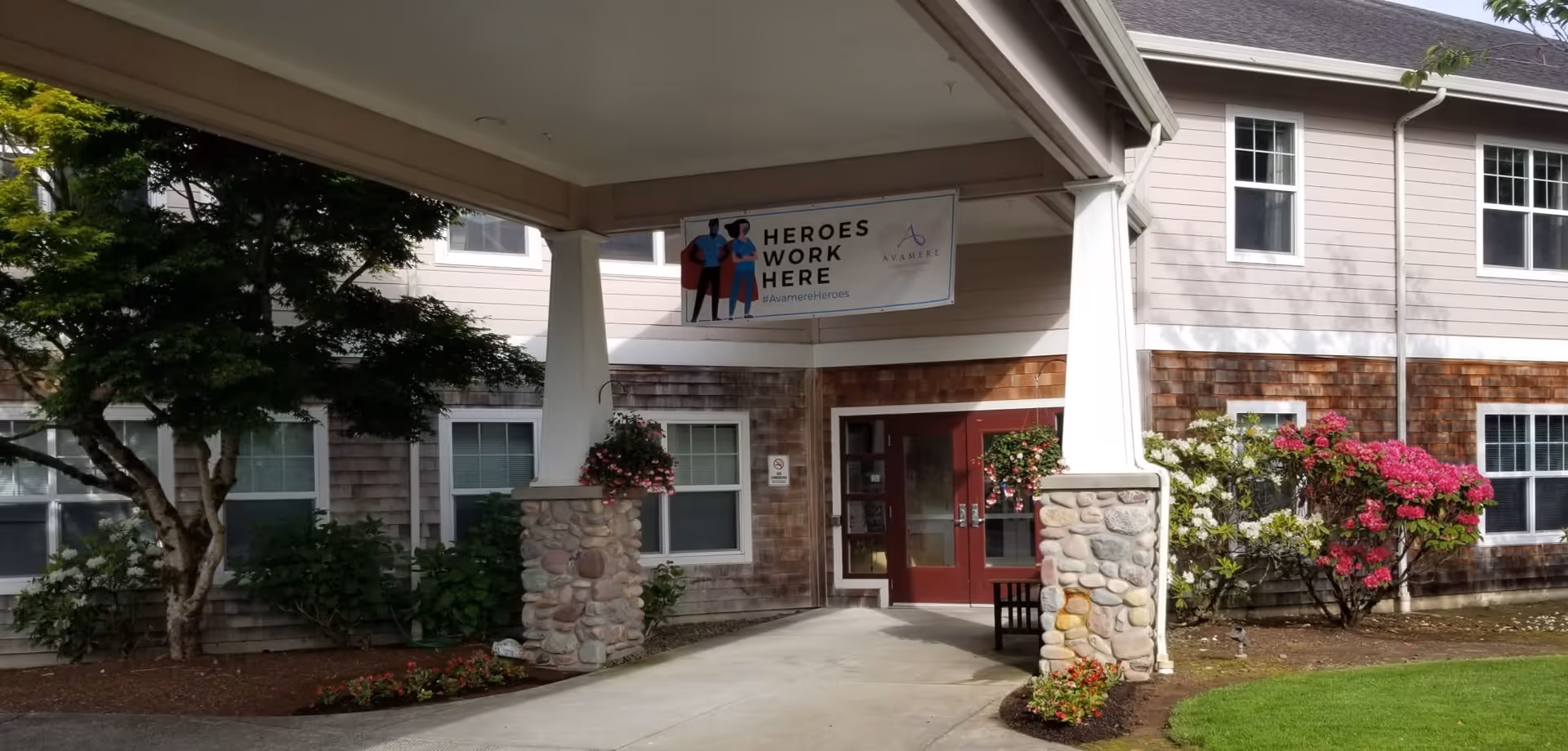 Entrance of a senior living facility with a covered walkway supported by stone pillars. There are hanging flower baskets and well-maintained landscaping with bushes and flowers. A banner above the entrance reads 'HEROES WORK HERE' with the Avamere logo and hashtag #AvamereHeroes.