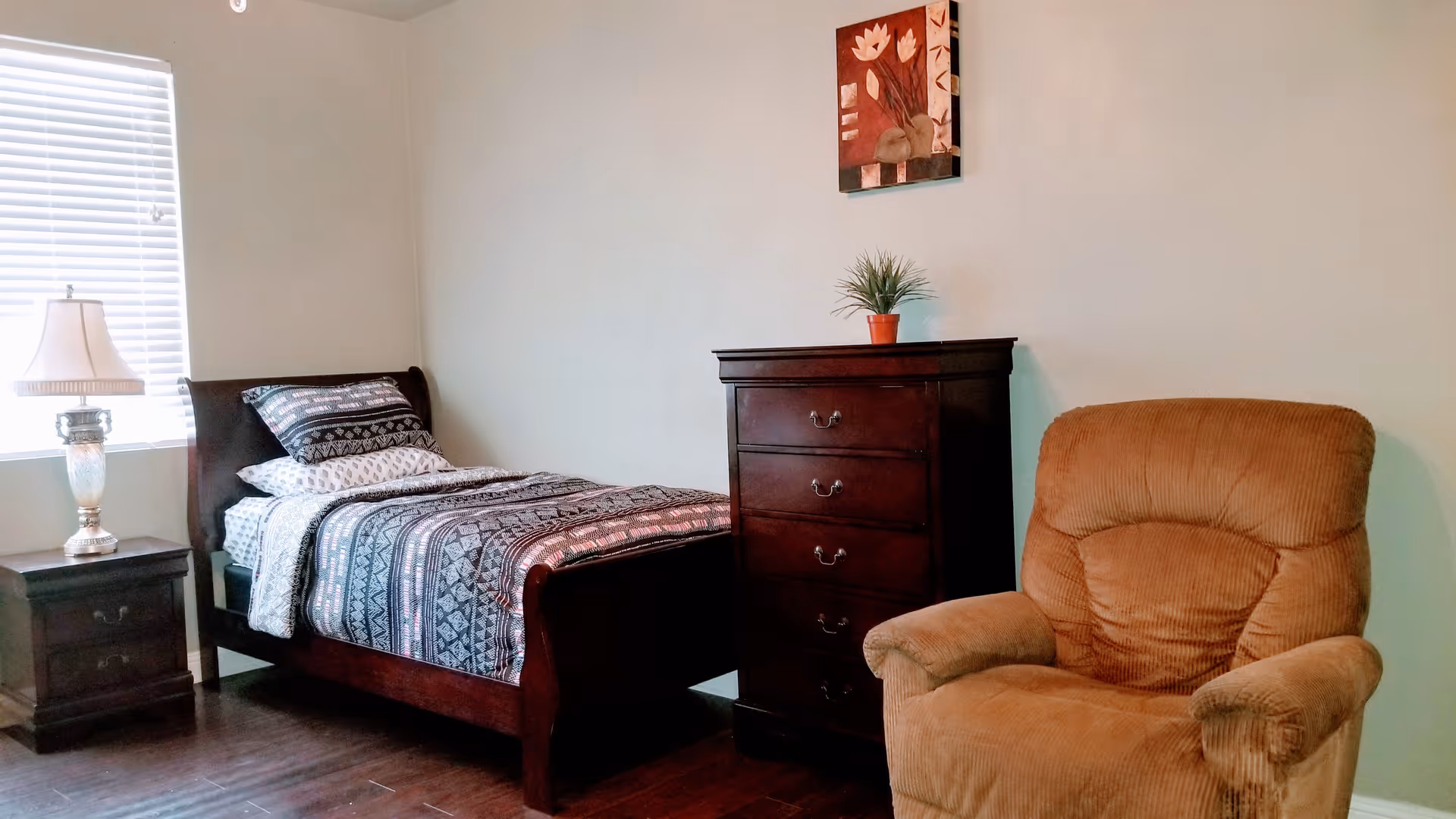 A cozy bedroom with a single bed featuring patterned bedding, a wooden nightstand with a lamp, a tall wooden dresser with a small potted plant on top, a comfortable brown armchair, and a window with blinds letting in natural light. A decorative painting hangs on the wall above the dresser.