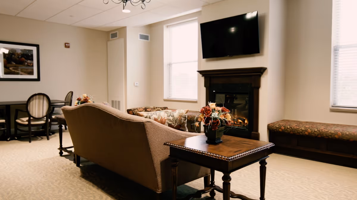 Cozy common living room with a sofa, side table with flowers, fireplace beneath a wall-mounted TV, and additional seating.