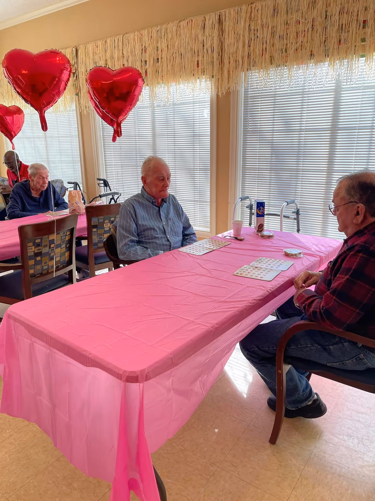 Two elderly men sitting across from each other at a table covered with a pink tablecloth, playing a game with cards. The room has large windows with blinds and is decorated with red heart-shaped balloons and a fringe garland along the top of the windows. Another elderly man is sitting at a nearby table in the background.