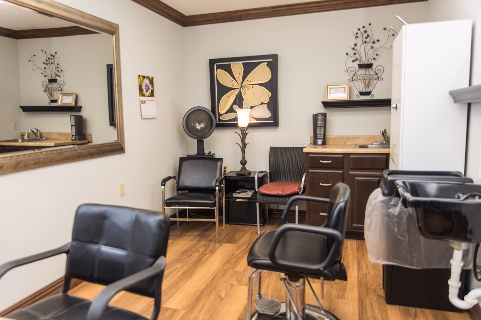 Interior of a small salon room with wooden flooring, featuring black salon chairs, a large wall mirror, a hair dryer, a lamp, a framed flower artwork, a small sink with countertop and cabinets, and decorative wall shelves with framed items and metal wall art.