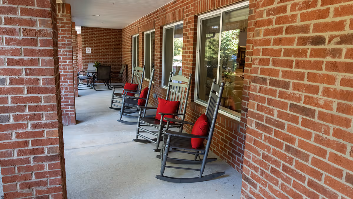 Covered outdoor patio area with four black rocking chairs each with a red cushion, positioned along a brick wall with windows. In the background, there is a small table with chairs and some greenery visible through the windows.