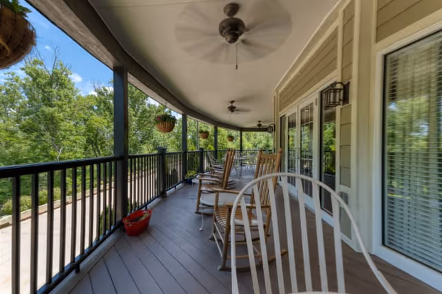 A covered outdoor porch with wooden rocking chairs and ceiling fans, overlooking a green wooded area. The porch has a railing, hanging flower pots, and glass doors leading inside the building.